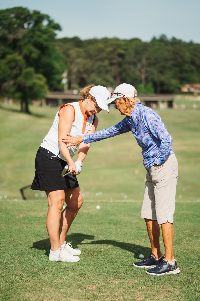 A golf instructor in a blue long-sleeve shirt assists a female student wearing a white top and black skirt with her swing technique on a sunny golf course.