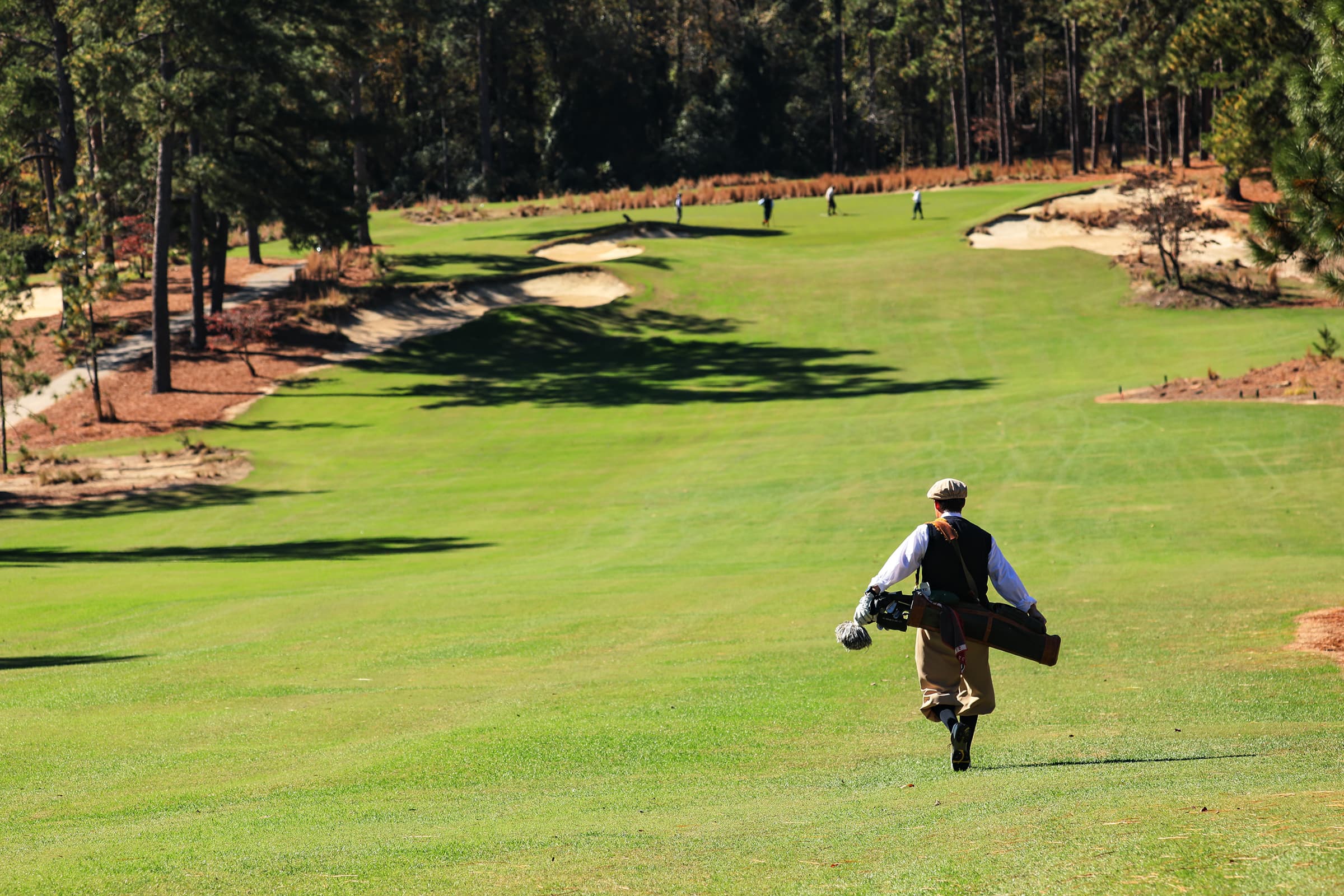 A person dressed in vintage golf attire walking down a well-maintained golf course. The background features lush green grass, trees, and sand traps. Other golfers are seen in the distance, enjoying their game.
