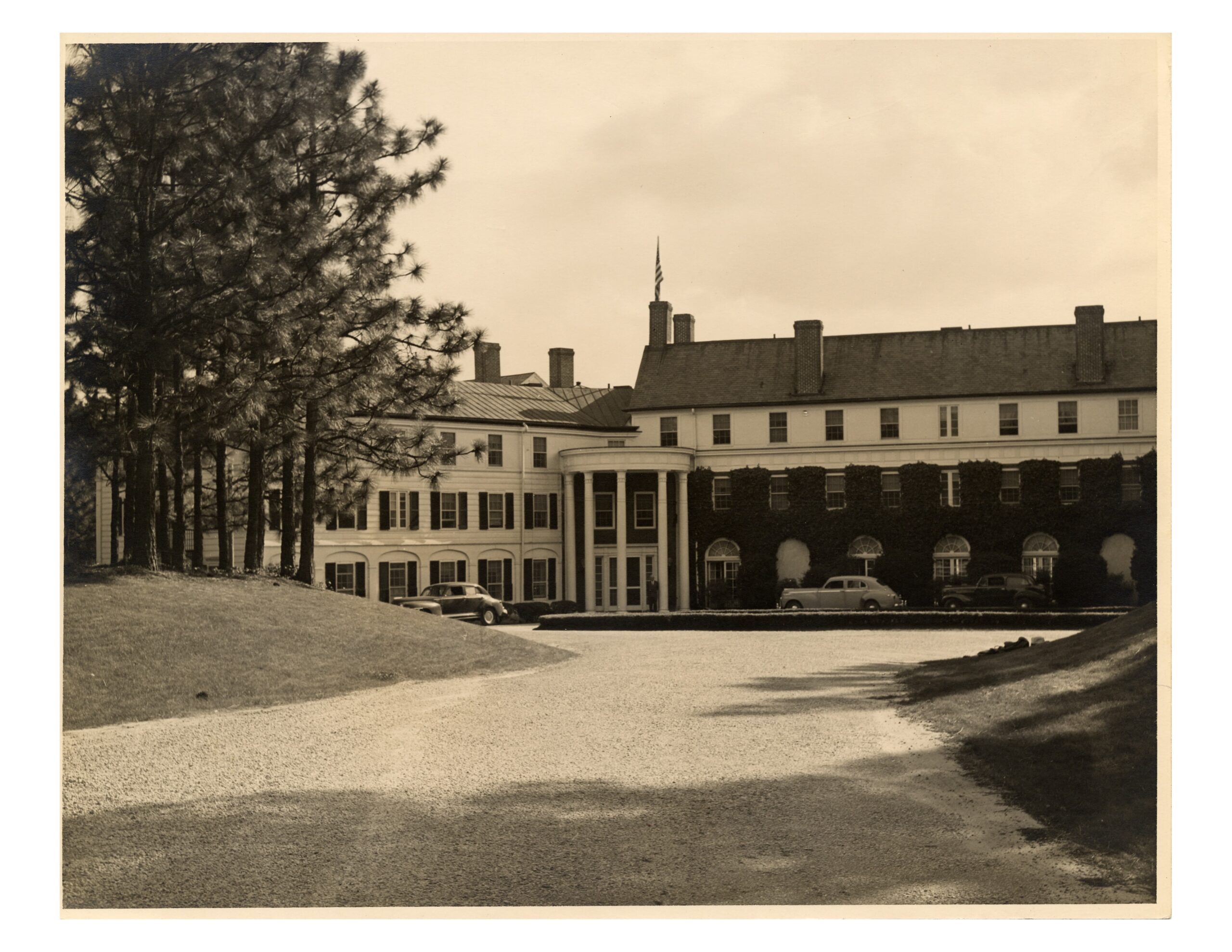 A historical hotel building featuring a central portico with columns, flanked by two wings. The structure is surrounded by trees and a gravel driveway leading to the entrance. Vintage cars are parked near the front.