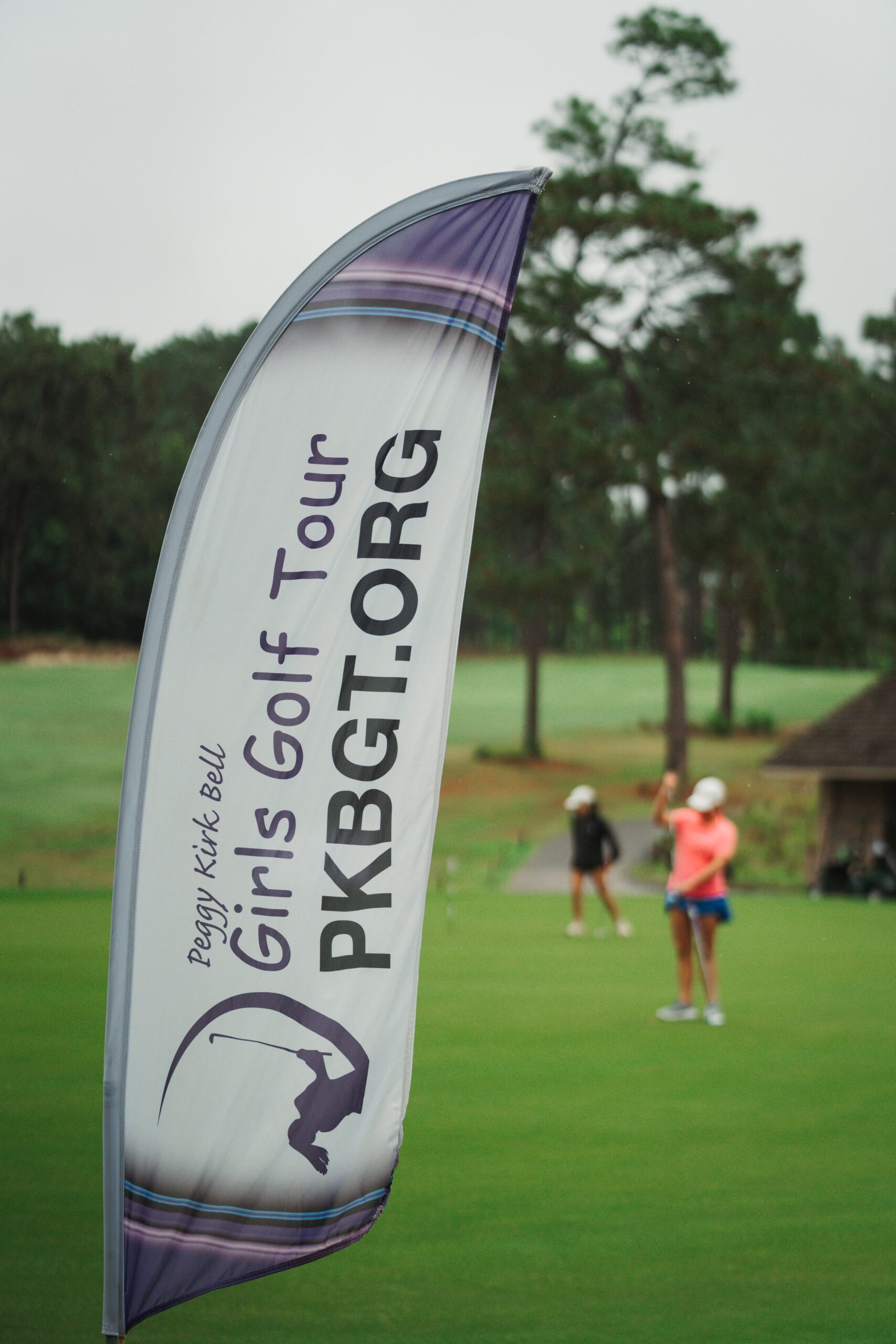 A banner for the Peggy Kirk Bell Girls Golf Tour is displayed prominently on the golf course. In the background, two female golfers are seen preparing to play. The setting features trees and a well-maintained lawn.