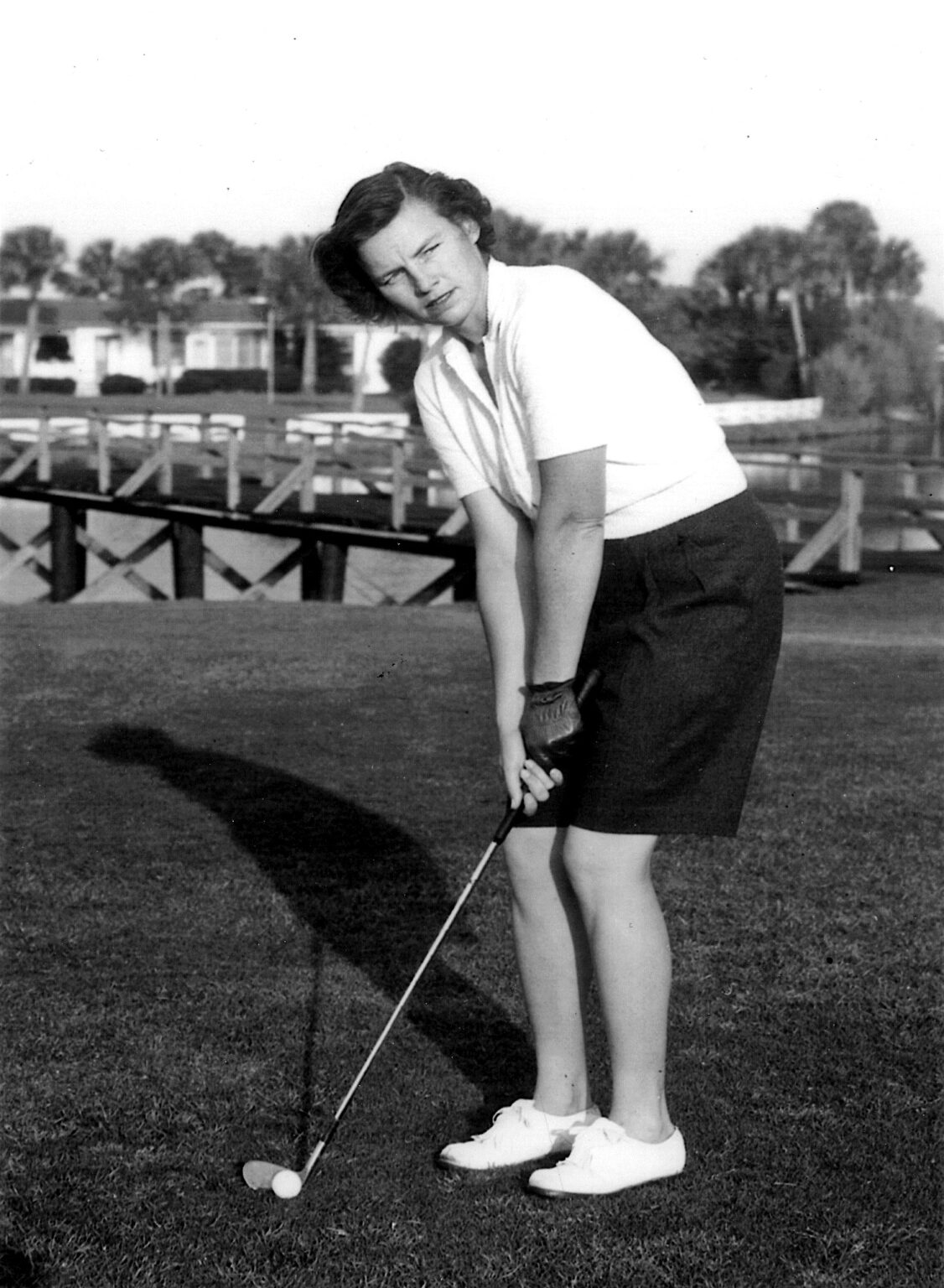 A woman preparing to putt on a golf course. She wears a white shirt, dark skirt, and white shoes. The background features palm trees and a wooden bridge. The image is in black and white.