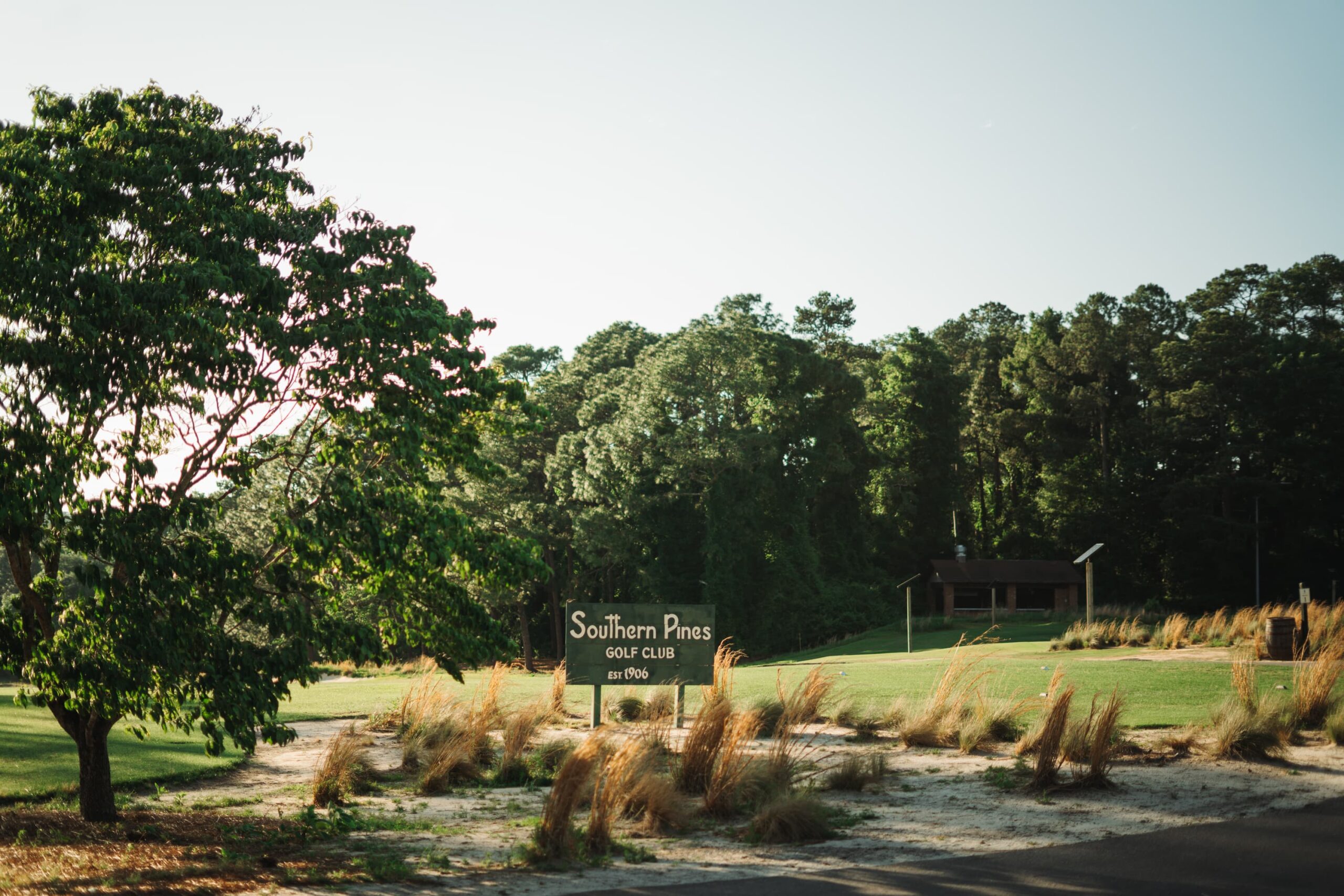 Entrance view of Southern Pines Golf Club, featuring a sign with the club's name. Lush greenery surrounds the area, including trees and tall grass. A wooden structure is visible in the background, suggesting additional facilities.