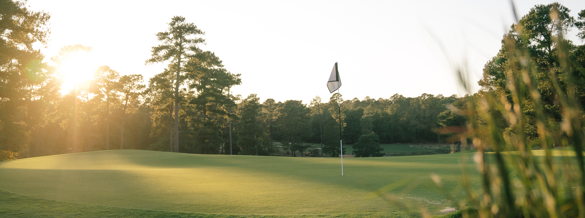 A golf course green in the early morning light, featuring a flagstick positioned on the putting surface. Surrounding the green are tall trees and a clear blue sky, with sunlight creating a warm glow over the landscape.