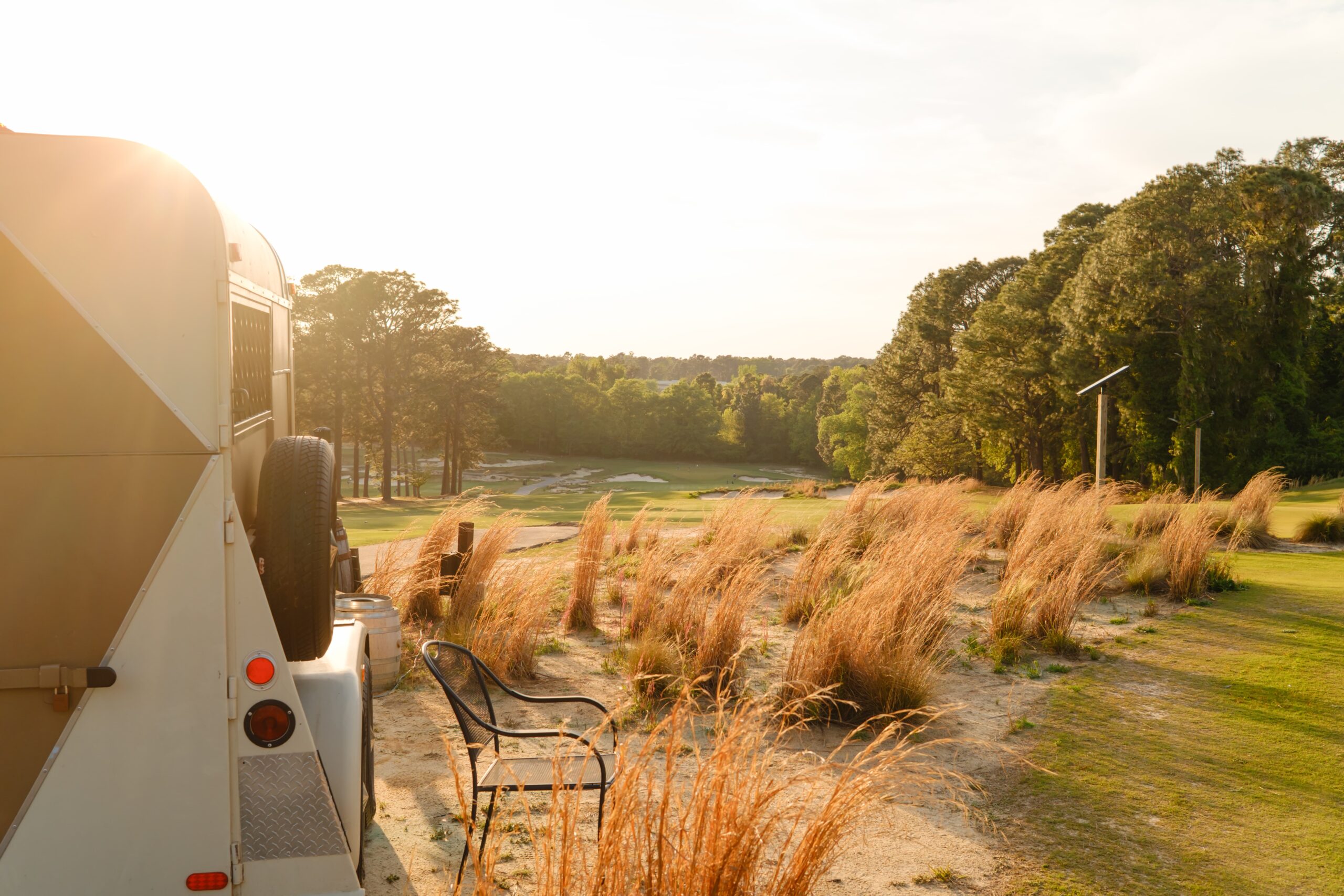 View of a golf course from the side of a camper, featuring tall grasses in the foreground. In the background, the course is visible with trees lining the edges and a setting sun illuminating the scene.