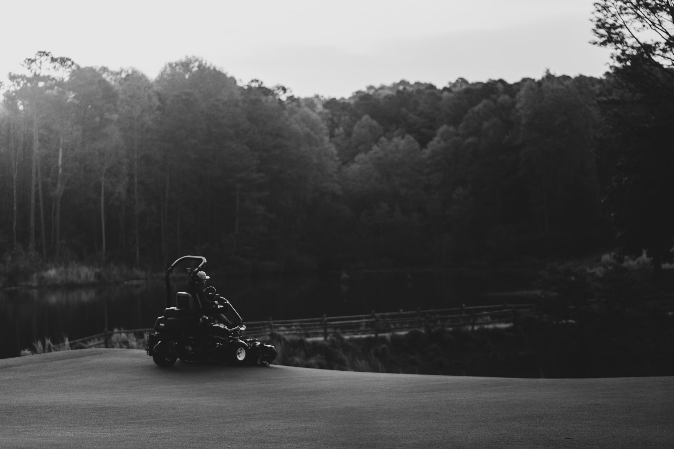 A golf course maintenance worker operates a riding mower on a grassy area near a lake. The setting includes trees in the background under a cloudy sky, presented in black and white.