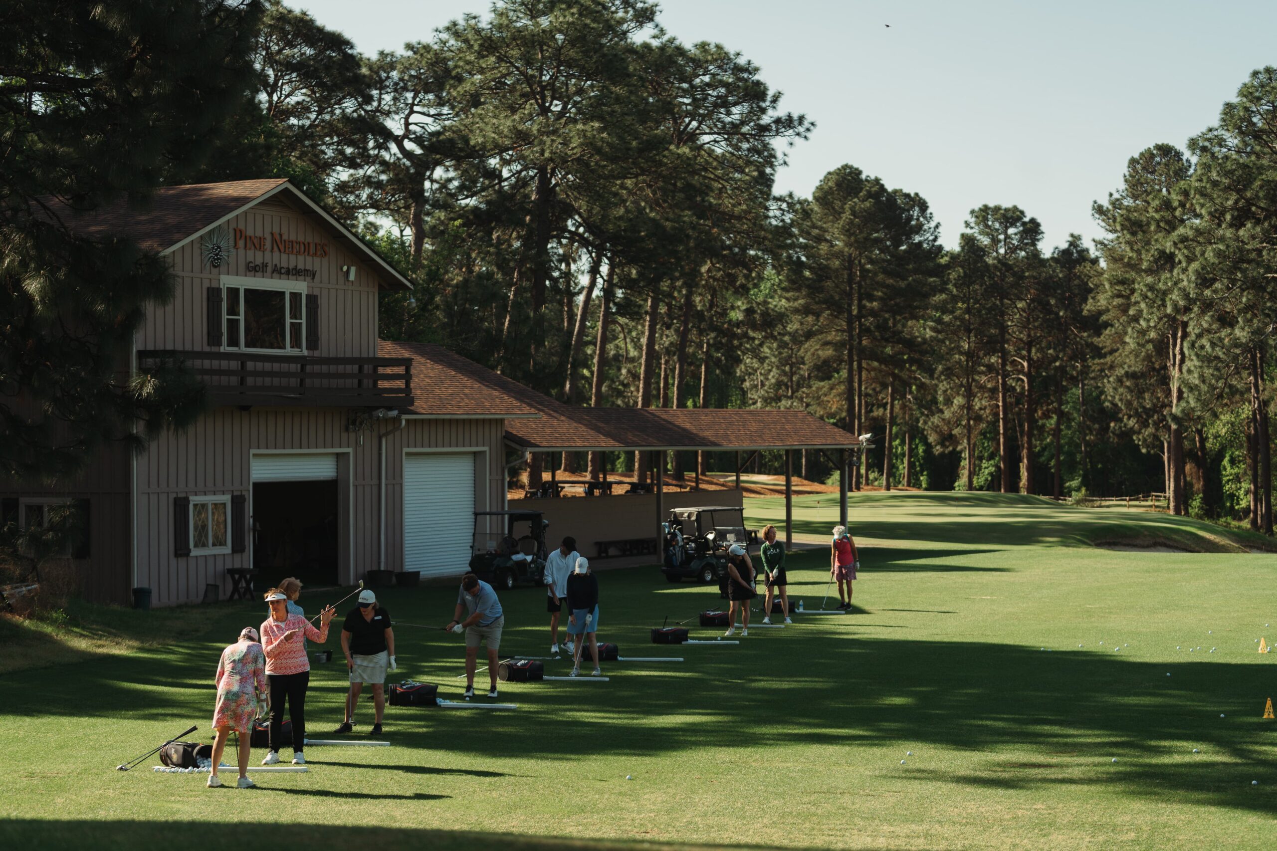 A view of a golf course with a training area, featuring several individuals practicing their swings. In the background, a wooden building serves as a clubhouse, surrounded by trees and green grass under a clear sky.