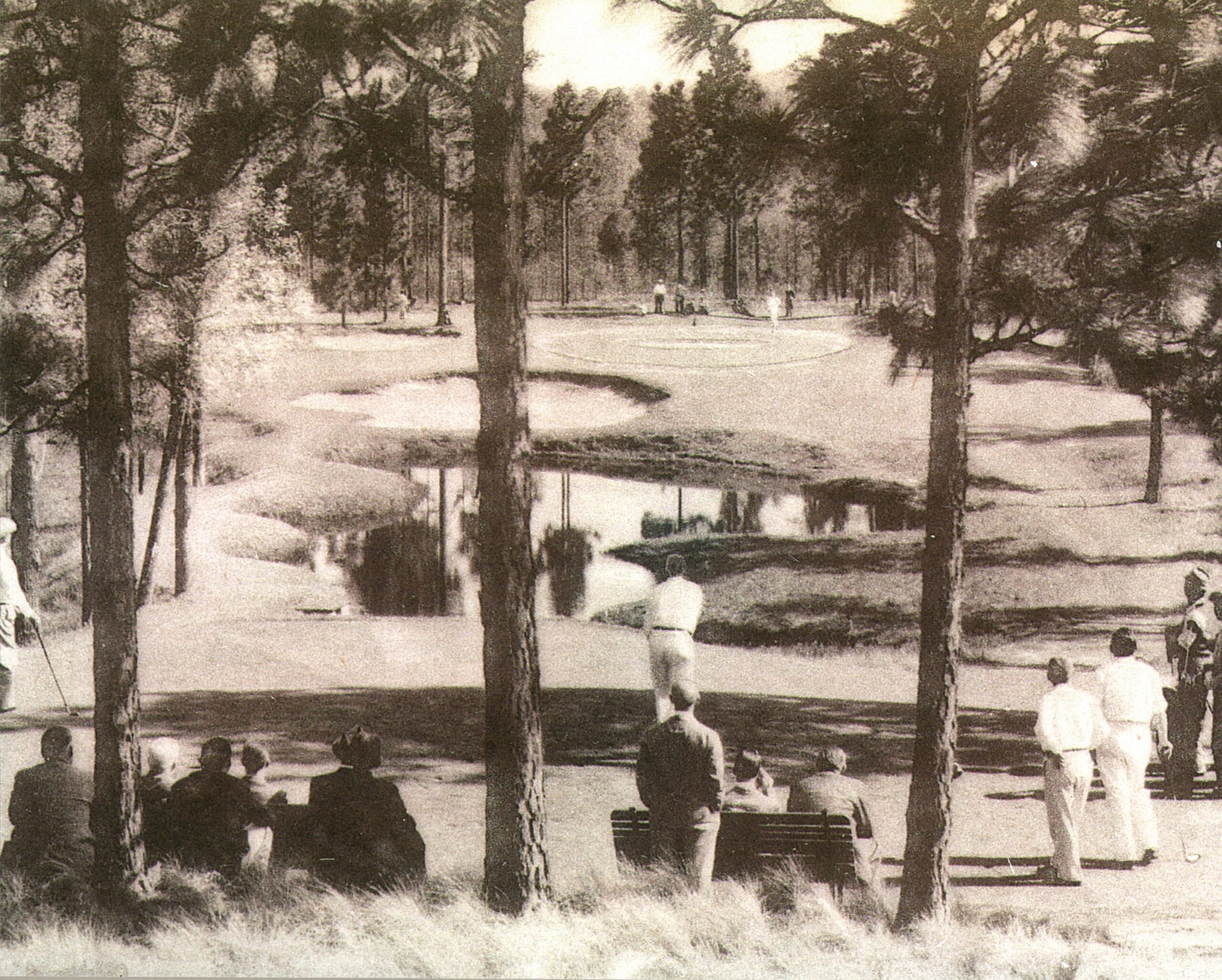 A vintage black-and-white photograph showing a golf course scene. Players are visible on the green, surrounded by trees. A group of spectators is seated on benches in the foreground, observing the game. A water hazard is present in the background.