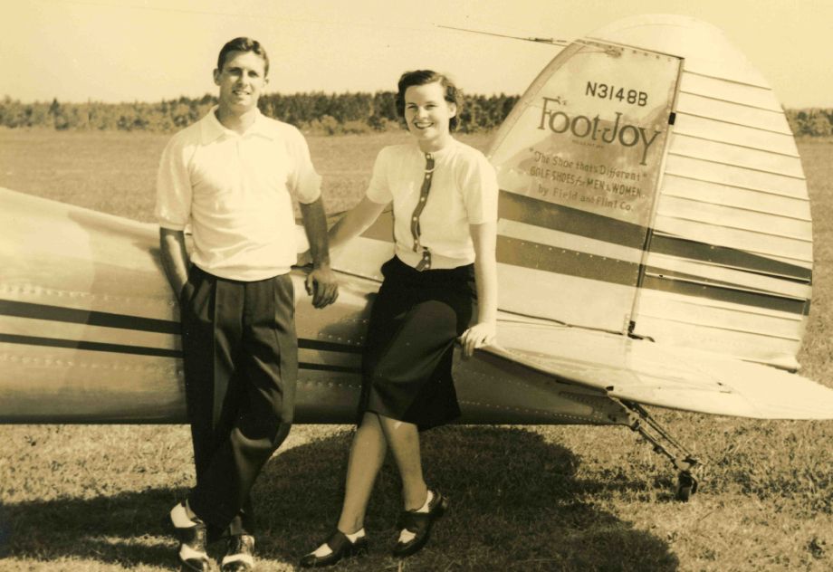 A man and a woman pose beside a vintage airplane. The man, dressed in a white polo shirt and dark pants, stands with his arm around the woman's shoulder. She wears a light blouse and a dark skirt. The background features open fields and trees.