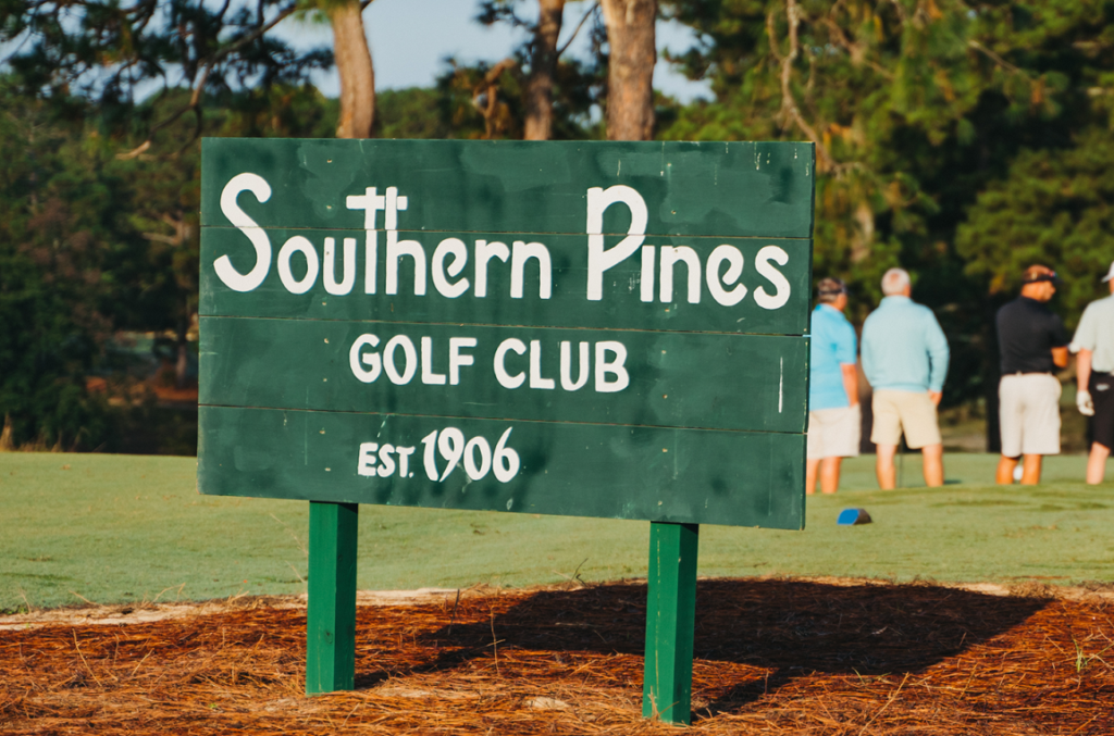 A green wooden sign for Southern Pines Golf Club displaying the text 'Southern Pines GOLF CLUB' and 'EST. 1906.' In the background, several people are gathered on a golf course under a clear blue sky.